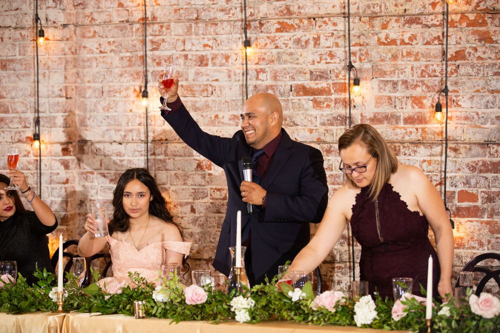 A celebration at a wedding reception with speakers at a head table decorated with greenery against exposed brick walls.