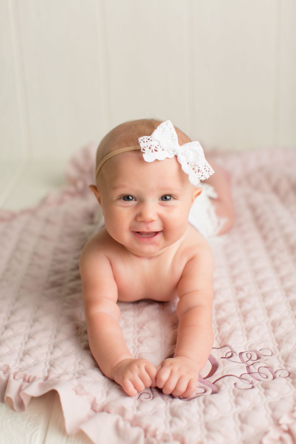 Baby lying on pink textured blanket wearing white lace bow headband.