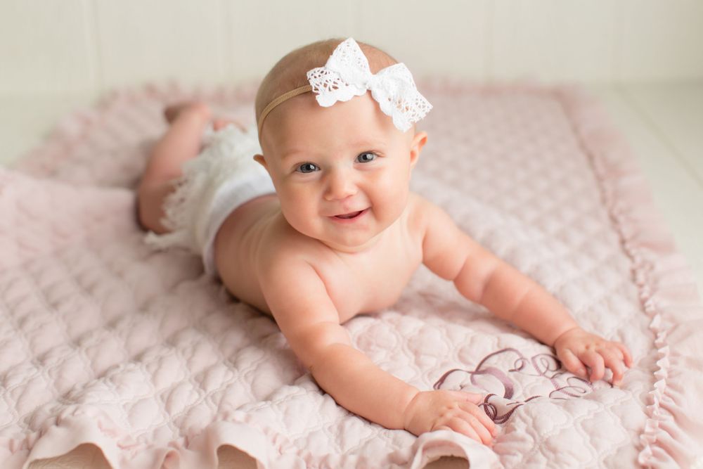 Smiling baby wearing white lace bow on soft pink blanket.