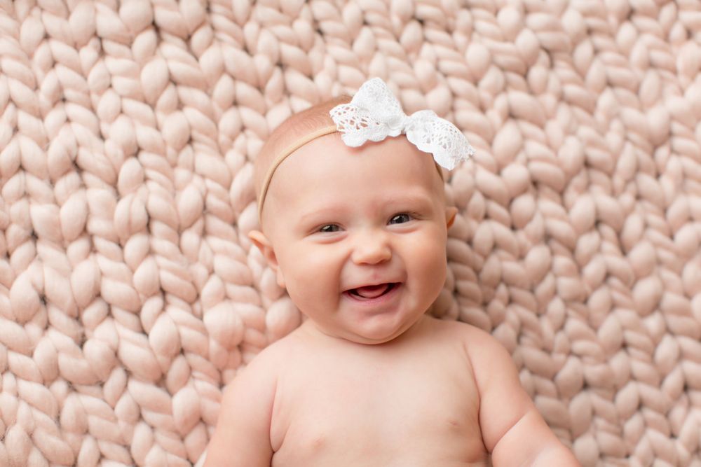 A smiling infant wearing a white bow headband lies on a chunky knit pink blanket.