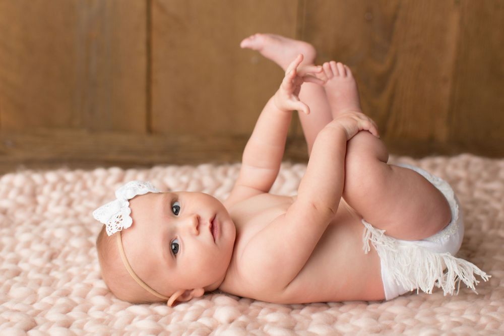 An infant in a white headband and diaper lies on back playing with their feet on a pink blanket.