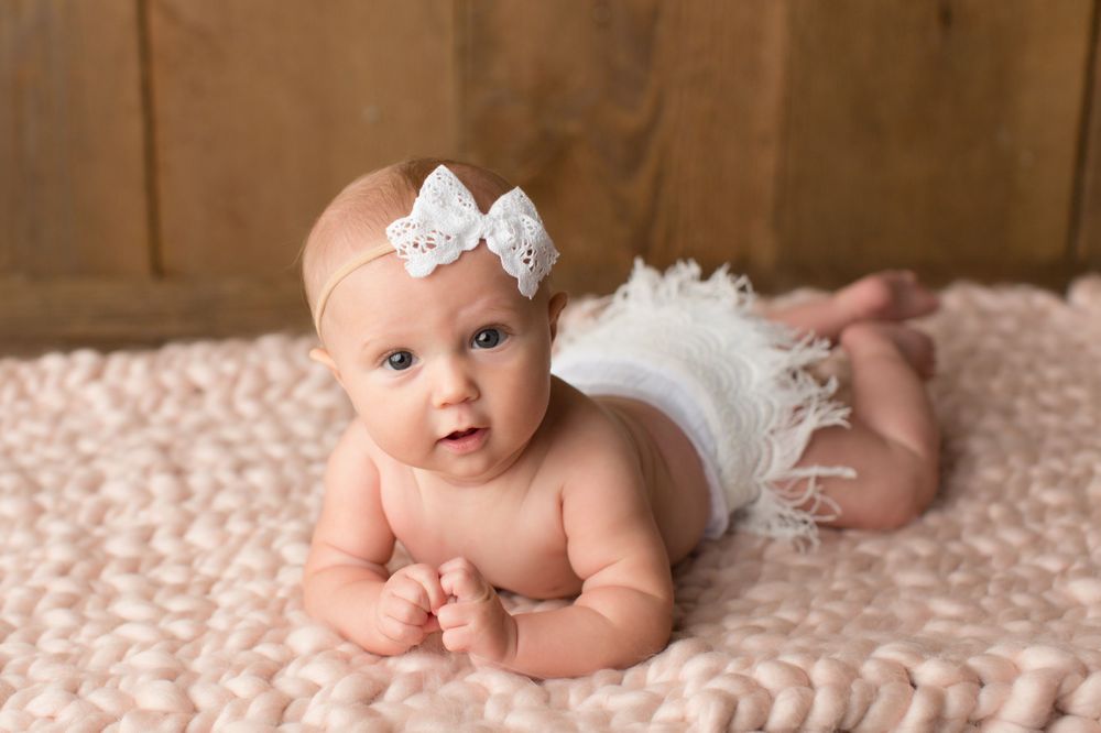 An infant wearing a white bow headband and ruffle diaper lies on stomach on textured blanket.