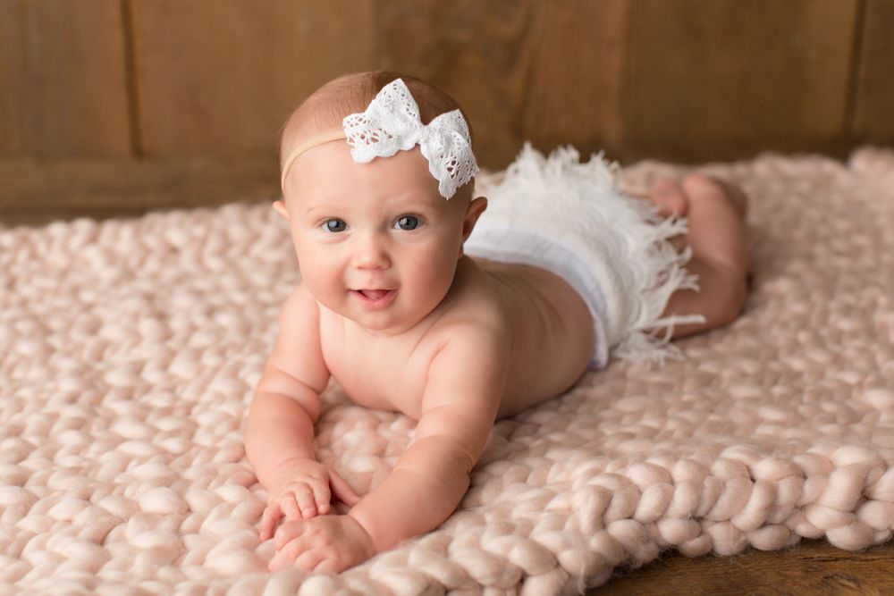An infant in white lace headband and diaper smiles while lying on chunky knit blanket.