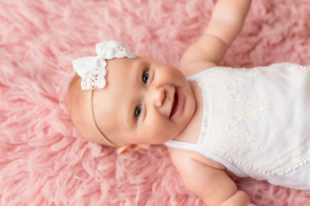 An infant in white lace outfit and bow headband smiles on pink textured blanket.