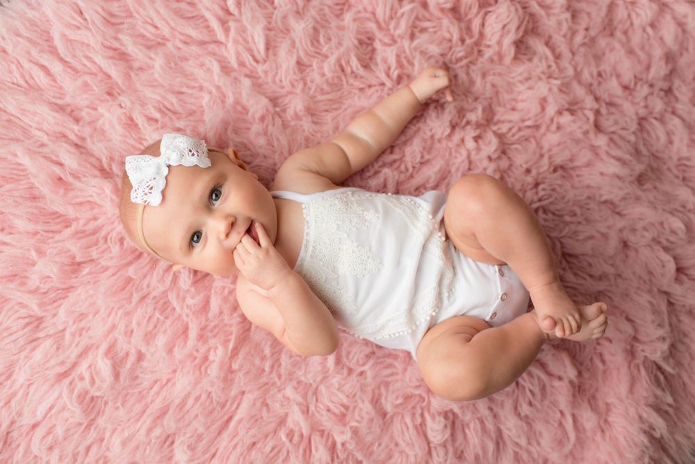 An infant wearing white outfit and bow headband lying on plush pink blanket.