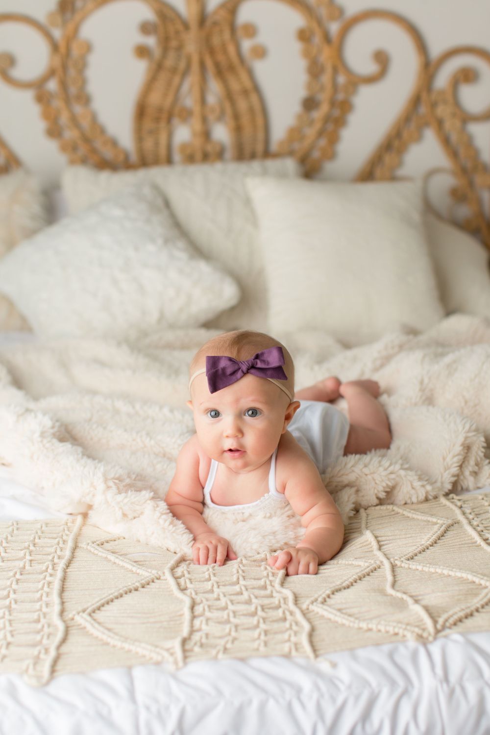 An infant in white outfit and purple bow rests on textured bedding with ornate headboard.