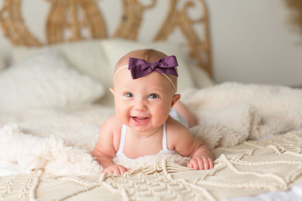 An infant wearing purple bow and white outfit smiles on cream textured bedding.