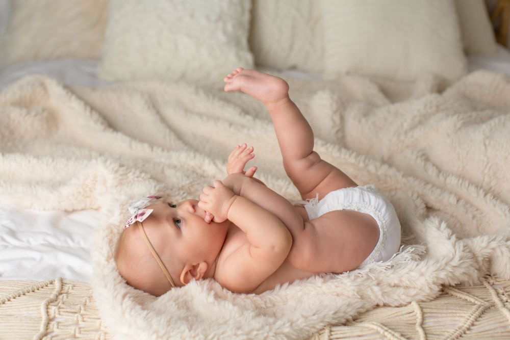 An infant wearing white headband plays while lying on textured cream bedding.