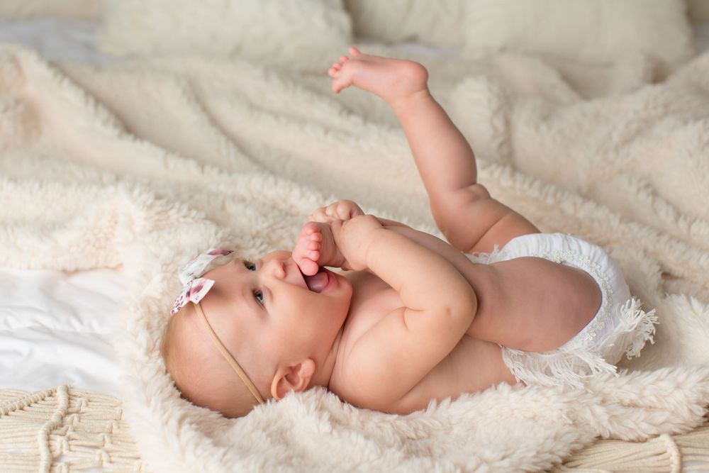 Infant in white diaper lies on soft cream blanket while stretching playfully.