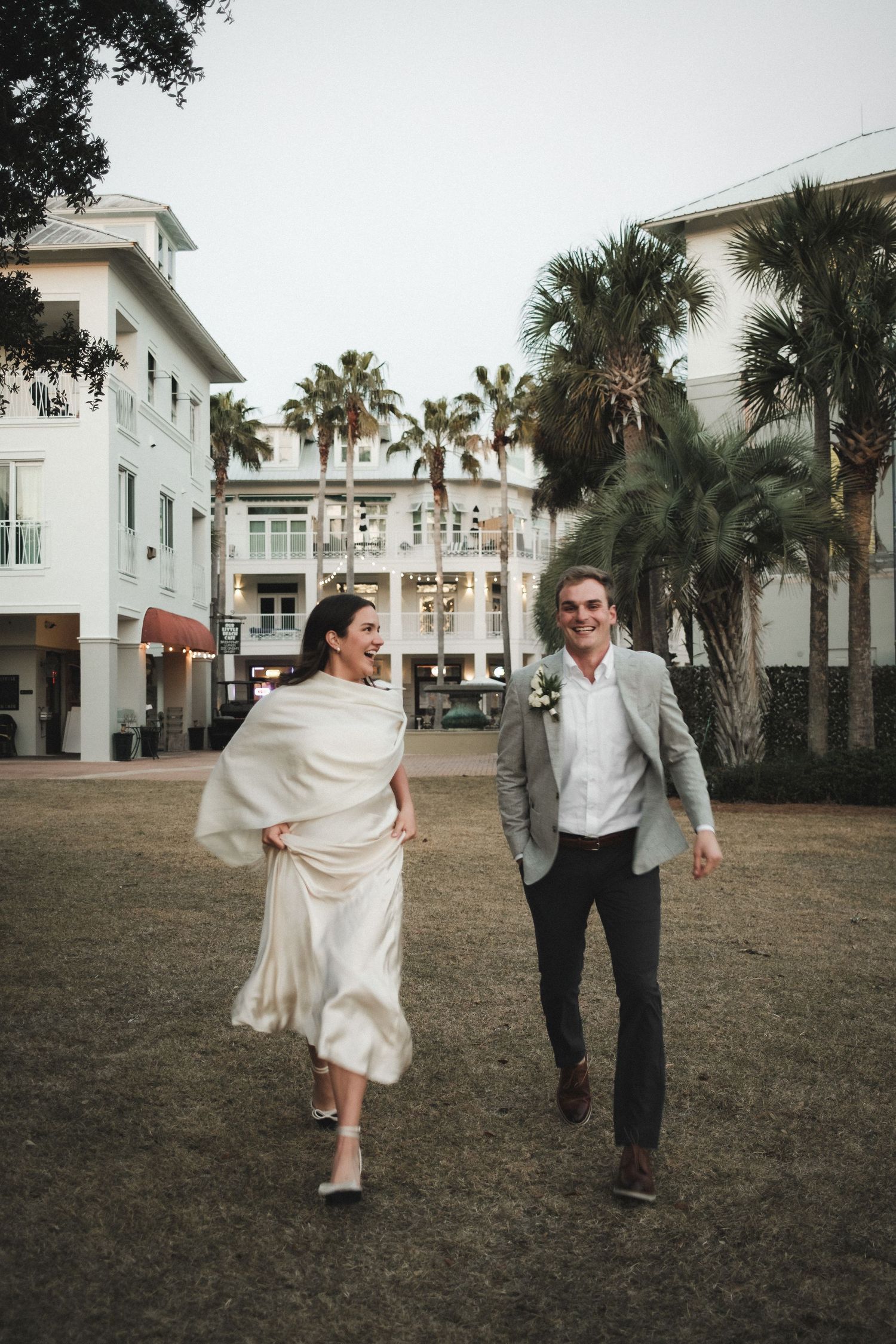 Couple walking and laughing together on grass with palm trees and white buildings in background.