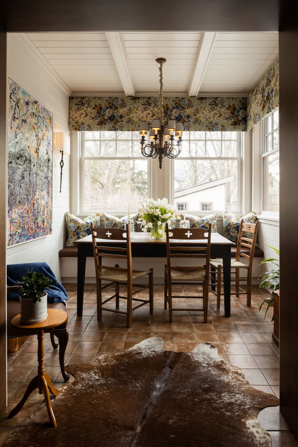 Cozy dining nook with chandelier, wooden table, and large windows featuring floral window treatments.
