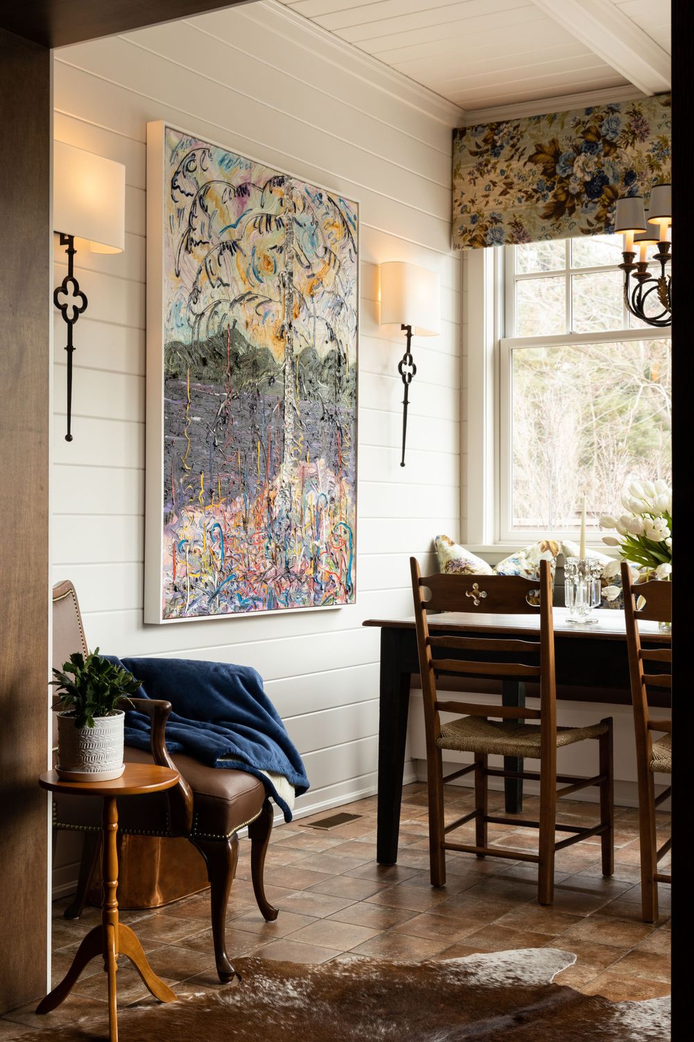 Rustic dining space with abstract wall art, wooden chairs, and cowhide rug on tile floor.