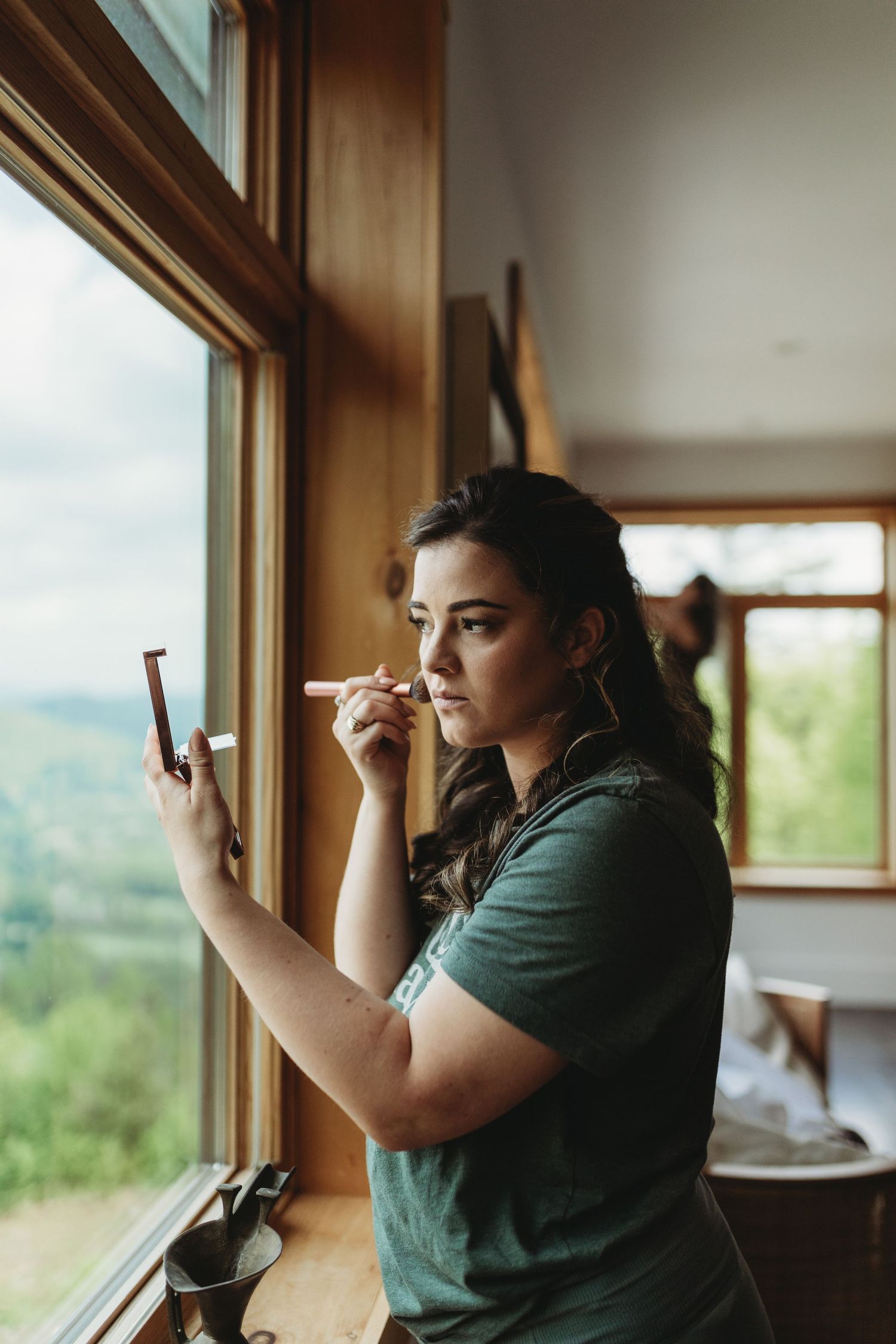 A person applies makeup while looking in a mirror by a window with mountain views.