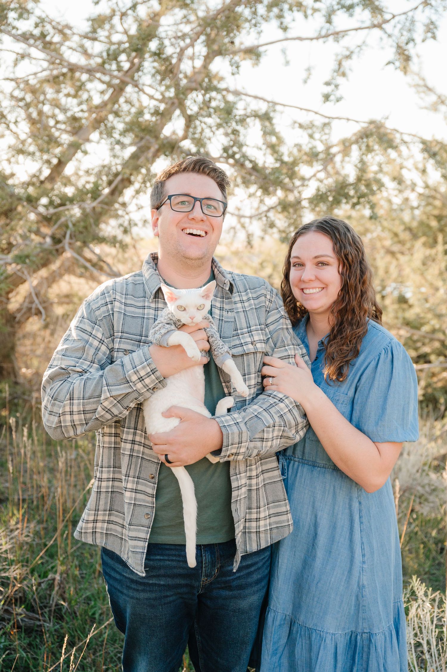Couple in casual attire holding white cats in outdoor natural setting.