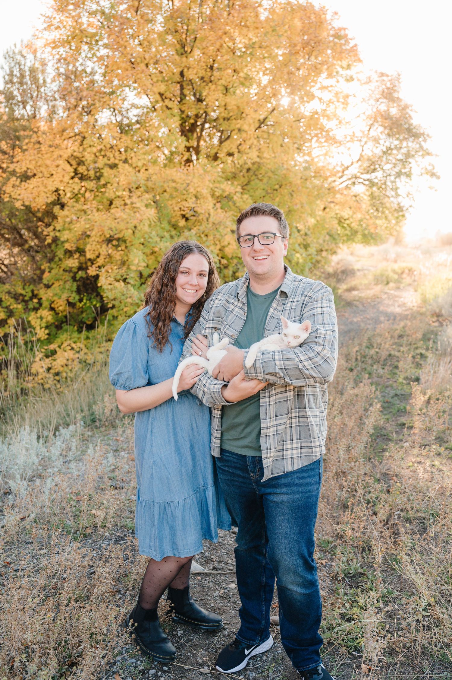 A couple poses outdoors in fall with golden trees, wearing casual clothing on a sunny autumn day.