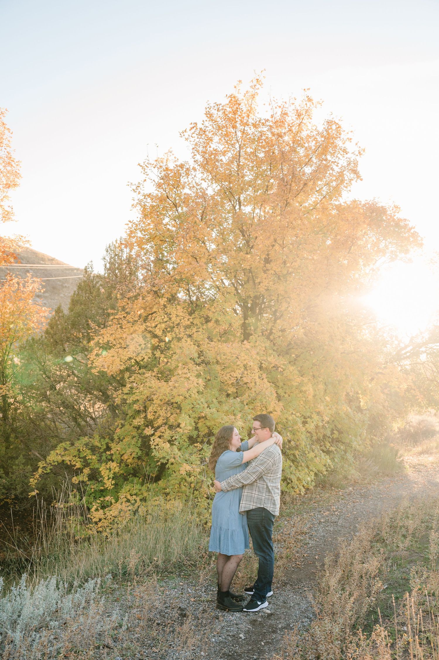 A romantic couple embraces on a trail with golden autumn foliage and sunlight in the background.