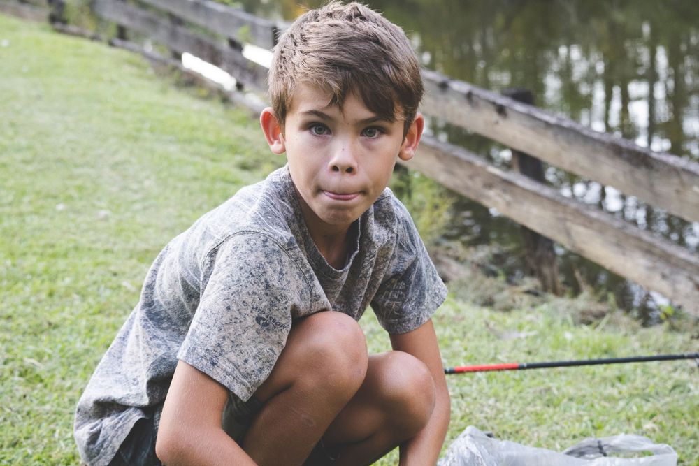 A pensive young child in a gray shirt squats outdoors on a grassy hillside. Fishing by pond.