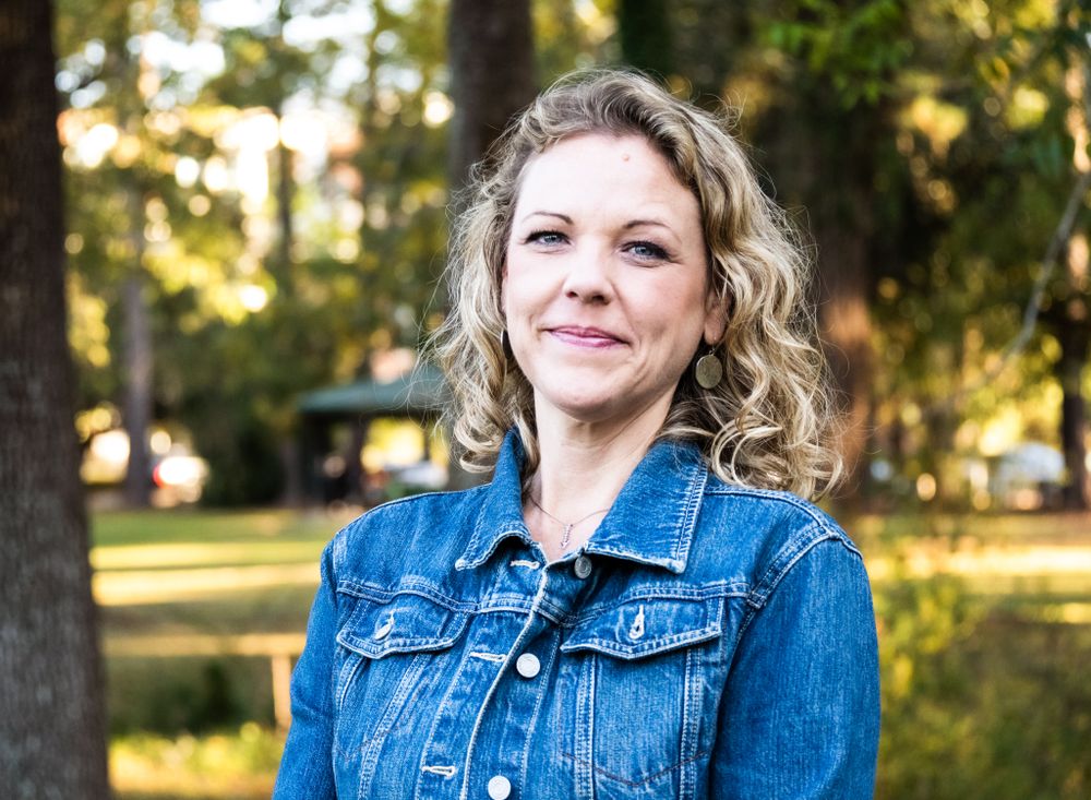 Woman with curly blonde hair wearing a denim jacket stands outdoors in a park with sunlit trees in the background.