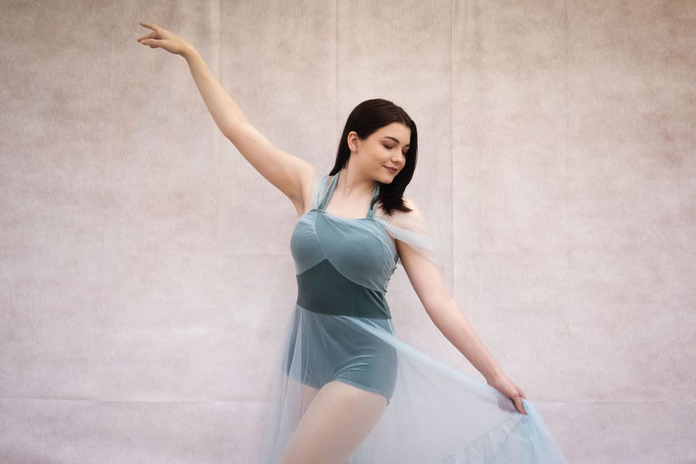 A dancer in a teal dress poses gracefully with one arm raised against a textured wall.