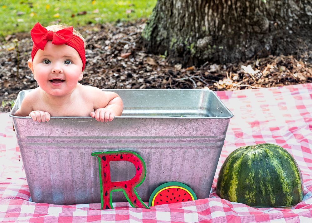 A small child sits in a galvanized metal tub next to a watermelon and red letter R on a picnic blanket outdoors. Baby photography.