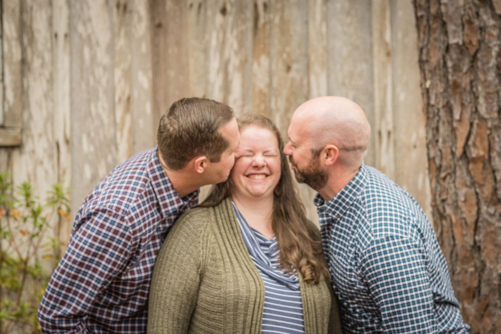 Three people share a joyful moment together outdoors against a rustic wooden wall backdrop.