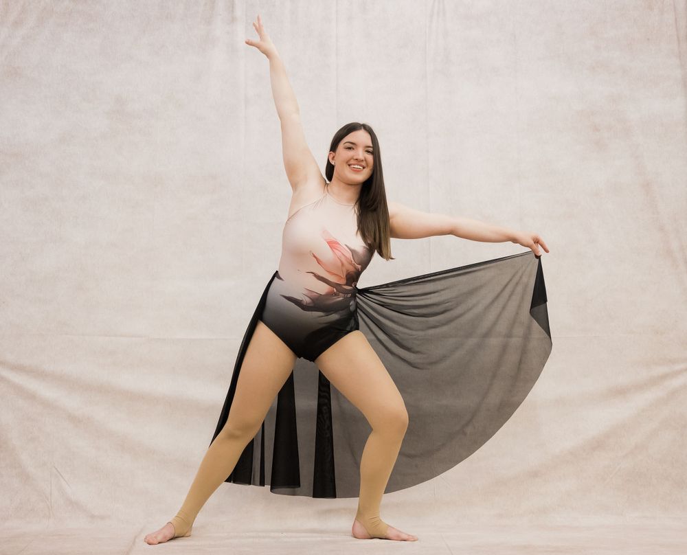 A dancer in a pink top and flowing gray skirt performs an expressive pose against a light backdrop.