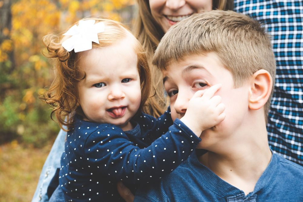 A young child in a polka dot dress and an older child make silly faces together with autumn foliage in the background.