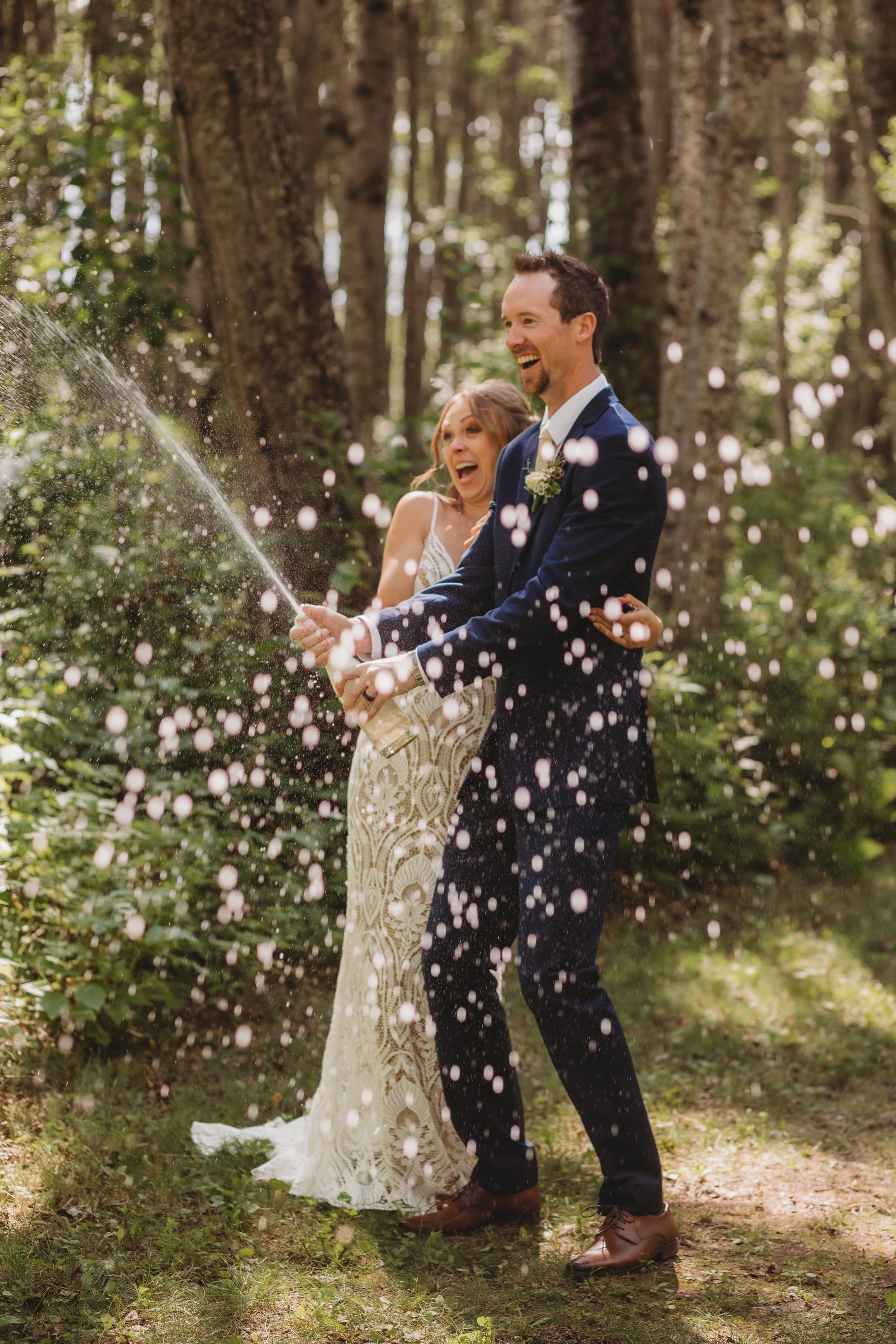 A joyful wedding couple sprays champagne in a wooded setting surrounded by sparkles and sunlight.