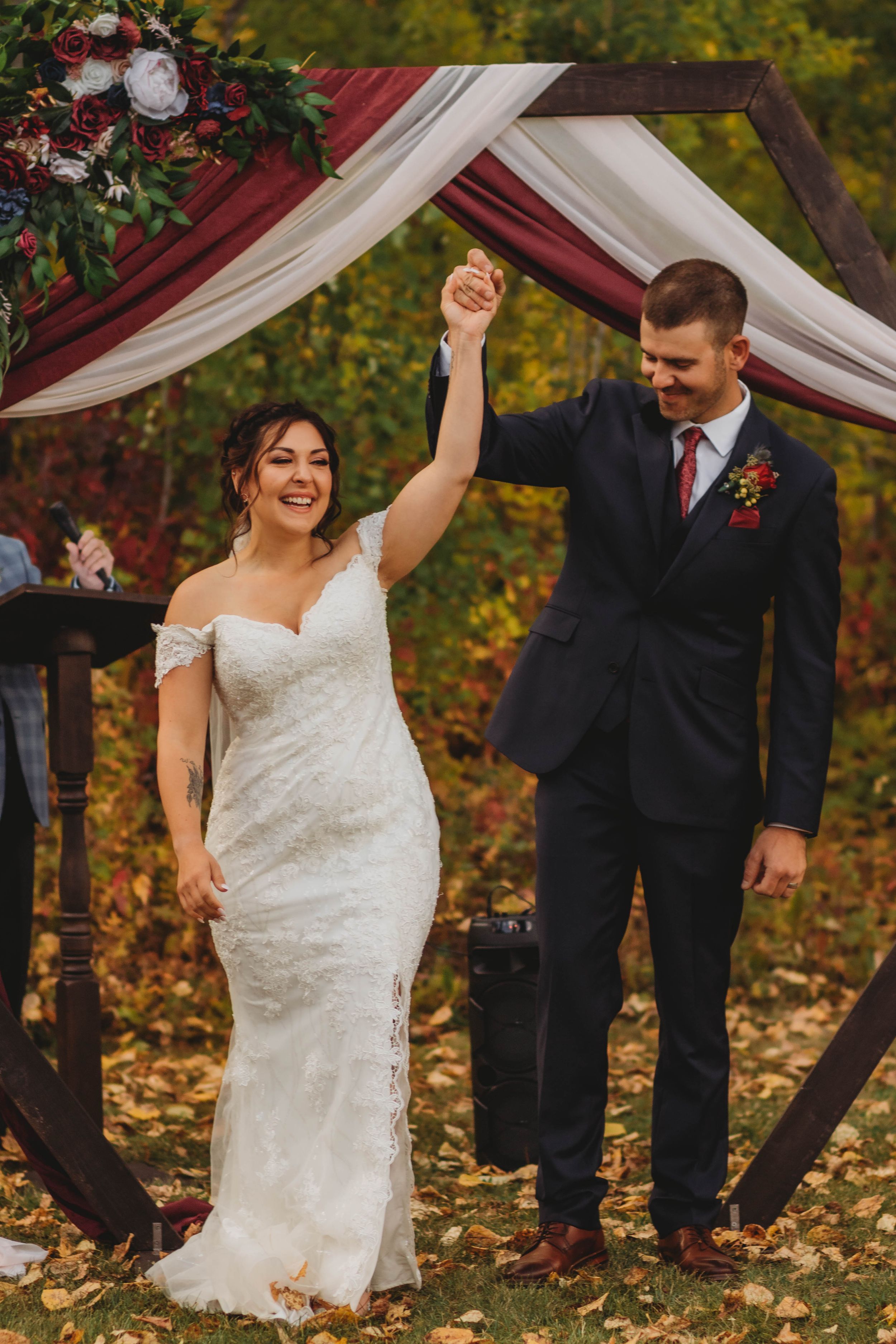 Newlyweds celebrate under a burgundy and white draped arch surrounded by fall foliage.
