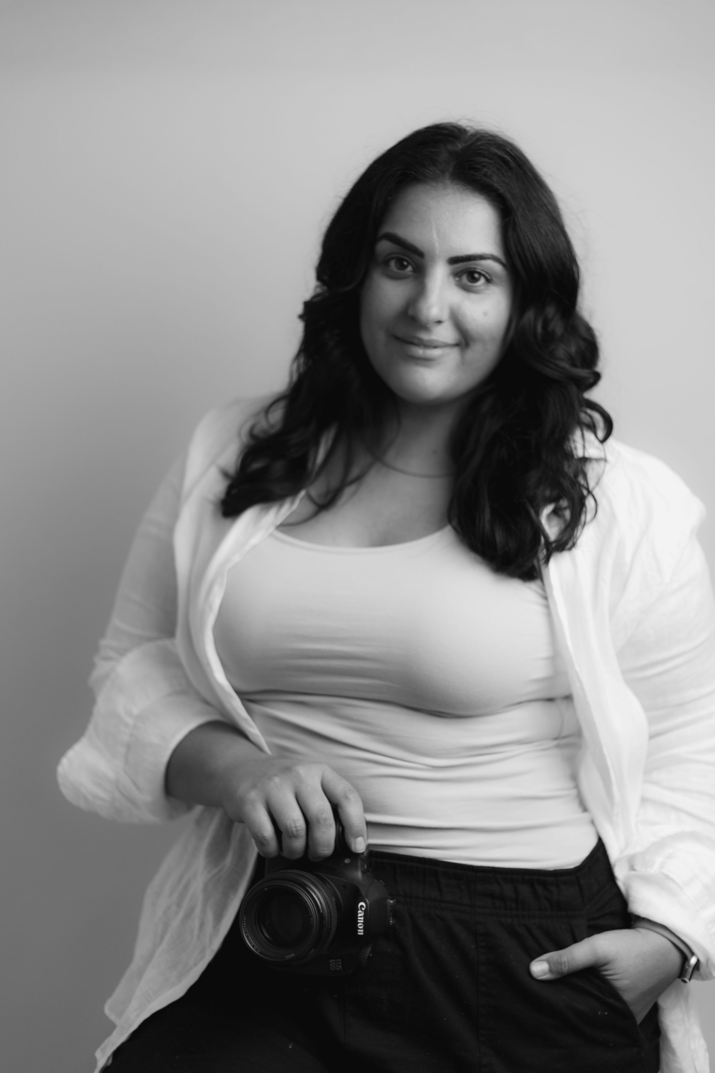 Portrait of photographer in white cardigan holding camera against grey background.