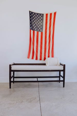 American flag hanging above a white bench against a light gray wall in a minimalist setting.