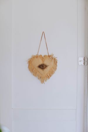 Decorative tan macrame heart hanging on a white wall.