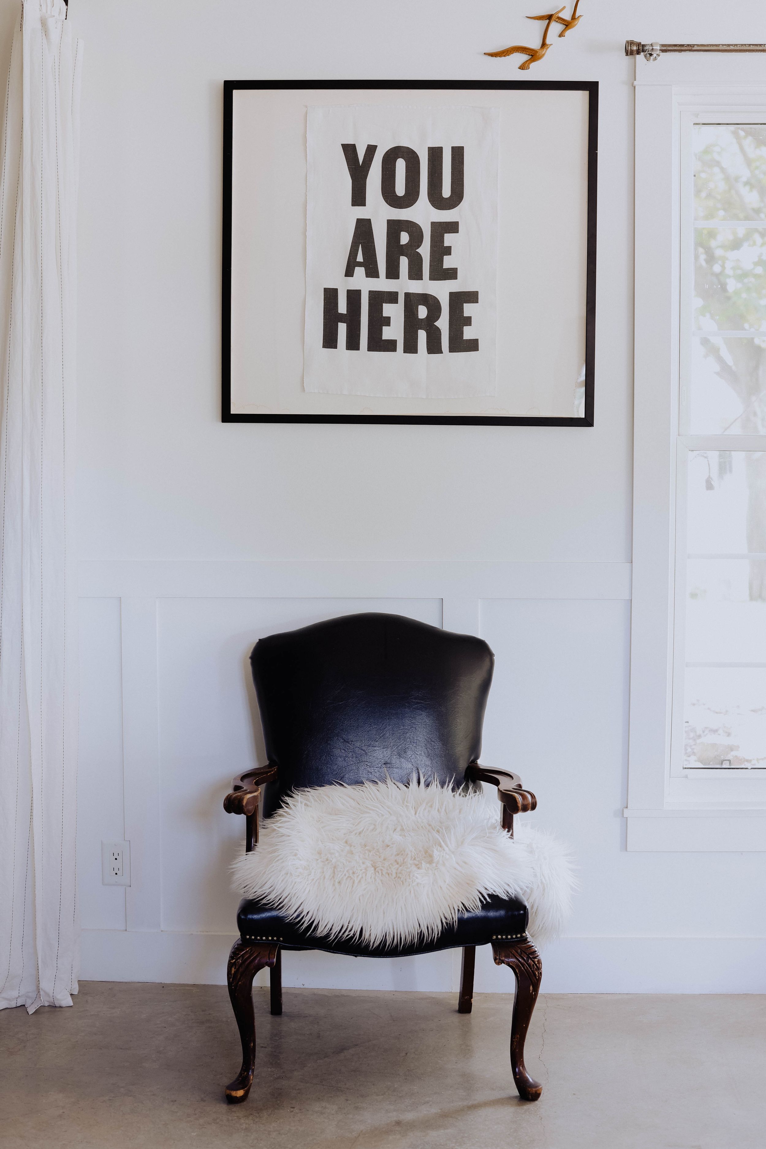 Vintage black chair with white fur cushion beneath 'YOU ARE HERE' wall art in minimalist room setting.