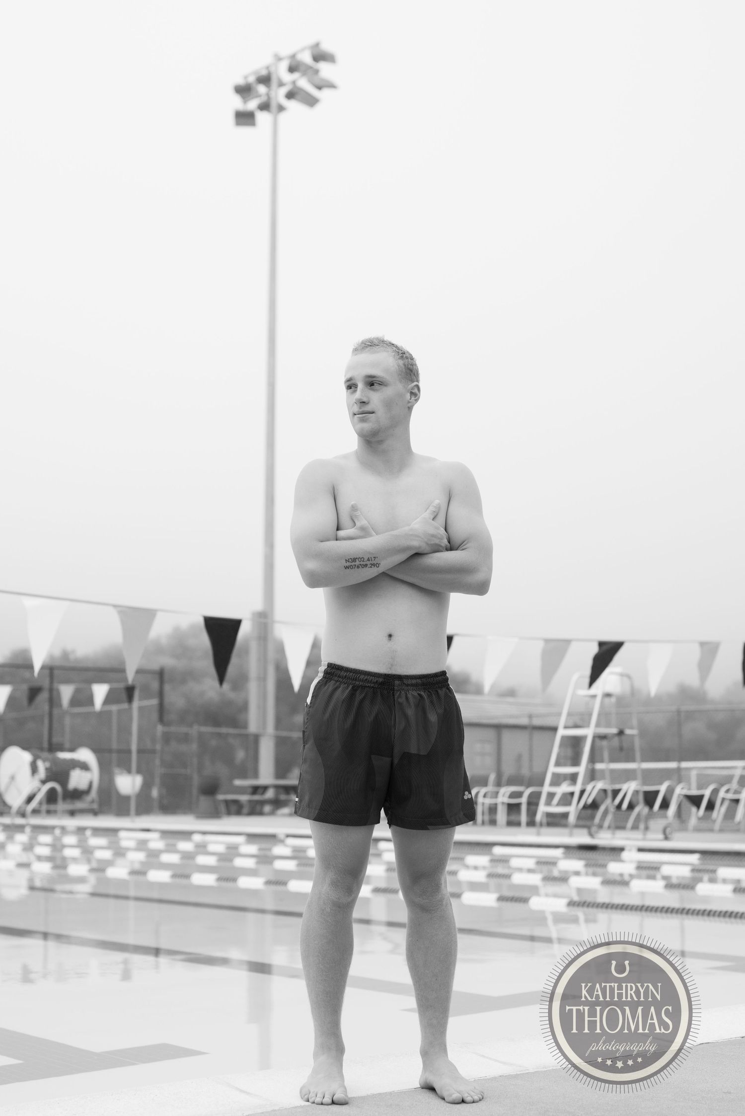 A swimmer stands poolside in swim shorts with a stadium light pole visible in the misty background.