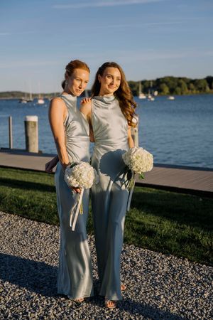 Two bridesmaids in elegant silver jumpsuits hold white bouquets while posing on a dock by a scenic waterfront.