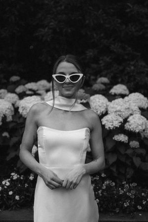 Black and white photo of someone in a white strapless dress and retro sunglasses posing in front of blooming hydrangea bushes.