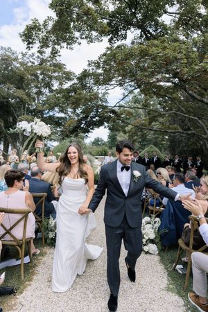 Wedding guests celebrate joyfully on a gravel path under trees at an outdoor reception ceremony.