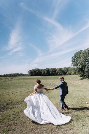 A couple in wedding attire spins together in a grassy meadow under a bright blue sky with wispy clouds.