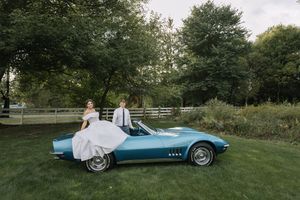 A classic blue Chevrolet Corvette convertible parked on a manicured lawn surrounded by trees and greenery.