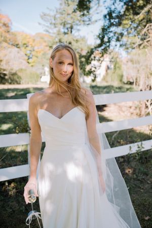 A bride in a classic white strapless wedding dress stands by a white fence on a sunny autumn day.