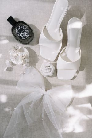 Elegant white bridal accessories including heels, tulle veil, and delicate details arranged on a light gray background.