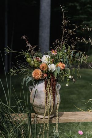 Rustic floral arrangement with dried flowers and greenery in a white ceramic vase displayed outdoors on wooden surface.