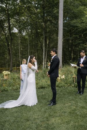 A wedding ceremony in a wooded outdoor setting with the couple and wedding party gathered on green grass.