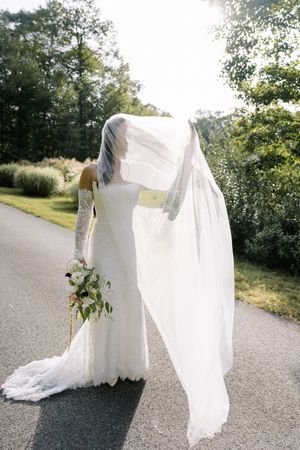 Elegant white wedding dress with flowing veil and floral bouquet photographed on a sunlit country road.