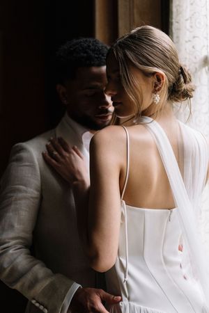 Intimate wedding portrait of couple embracing in soft window light, bride wearing elegant white dress with open back detail.