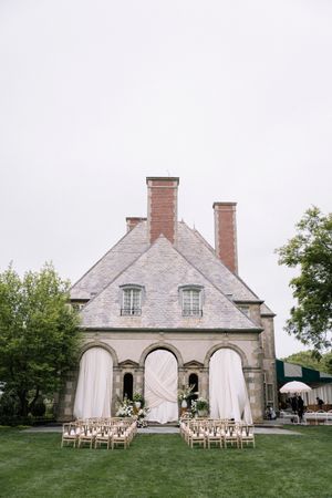 Elegant outdoor wedding ceremony setup with wooden chairs facing a stone building with three arched doorways and flowing curtains.