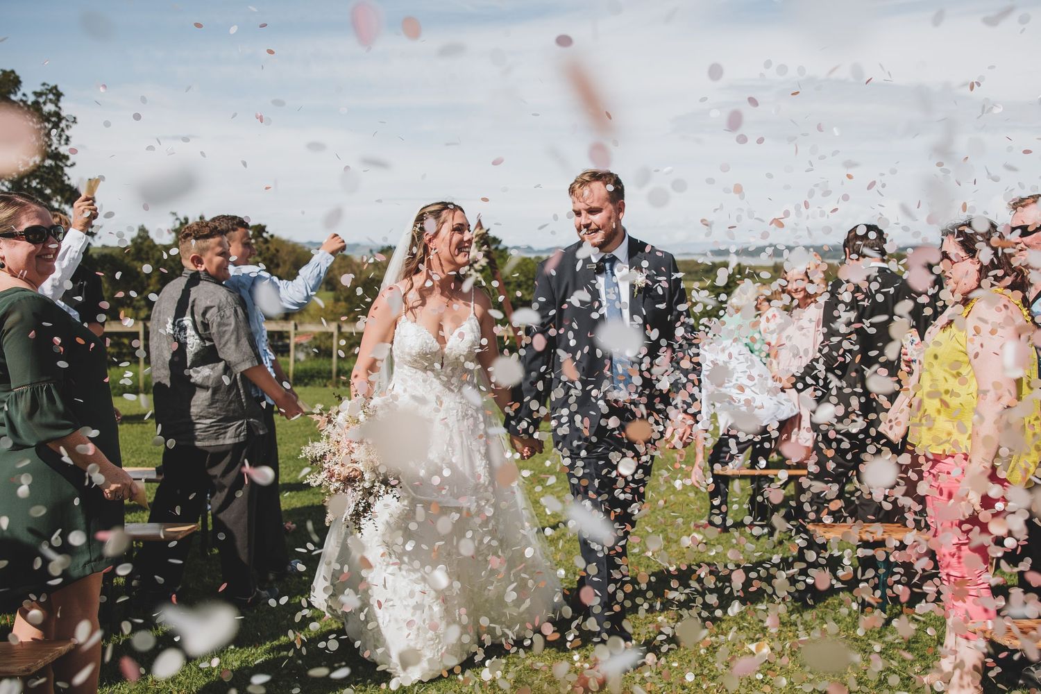 Wedding party celebrates outdoors with confetti flying through the air during a festive moment.