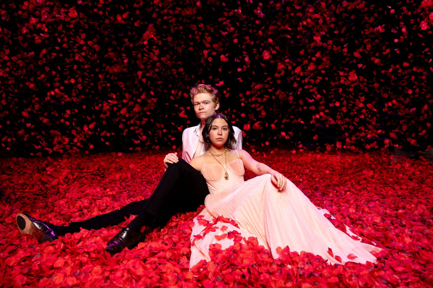 Romantic couple pose on red rose petals, with woman in pink gown and man in black attire.
