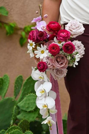 A floral arrangement featuring red ranunculus, white orchids, pink roses, and daisies against an adobe background with cacti.
