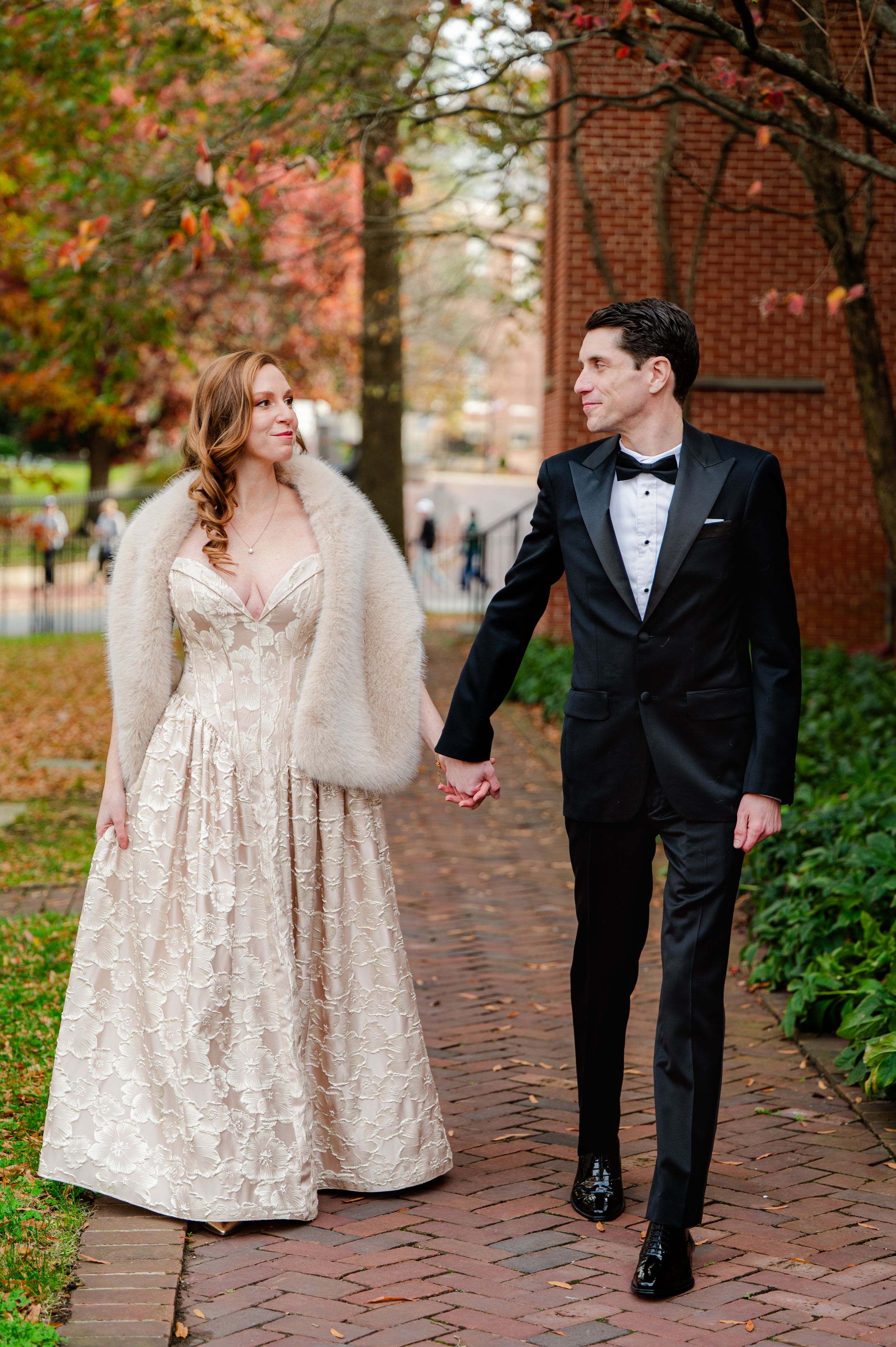 Bride in lace gown with fur stole and groom in black tuxedo walk hand-in-hand on brick path among autumn trees.